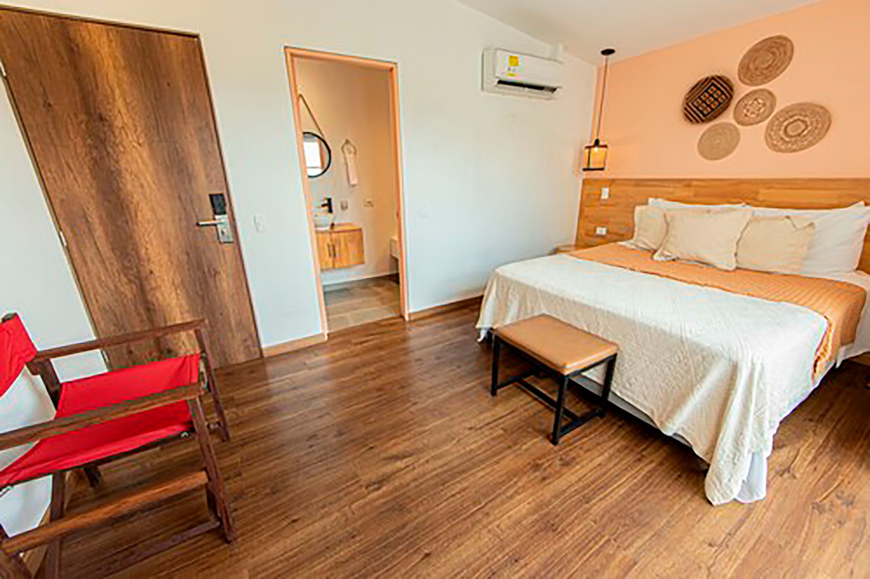 Une chambre avec un plancher en bois, un lit avec une literie blanche, un banc, une porte en bois et un climatiseur fixé au mur.