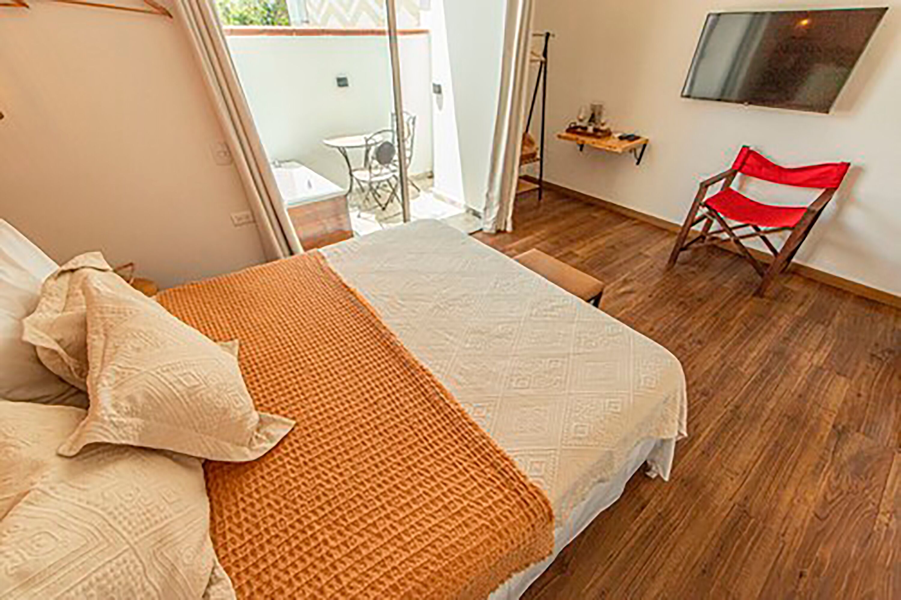 A bedroom with a bed, pillows, a blanket, a wooden floor, a red chair, a TV on the wall, and a small table with a plant.