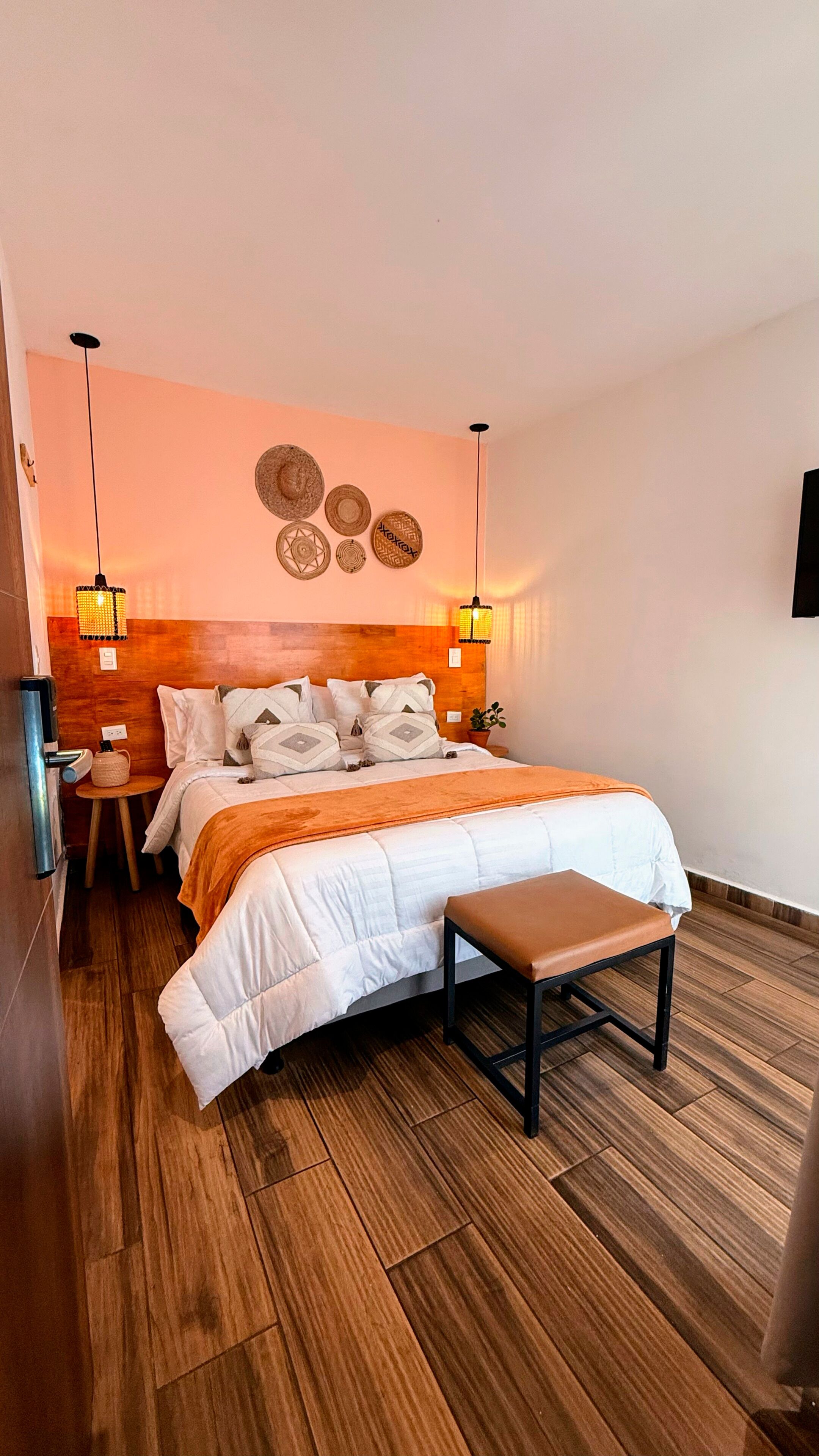 Et moderne hotelværelse med trægulv, en stor seng med hvid dyne, et trænatbord, et lille bord med lampe og et TV ophængt på væggen.
