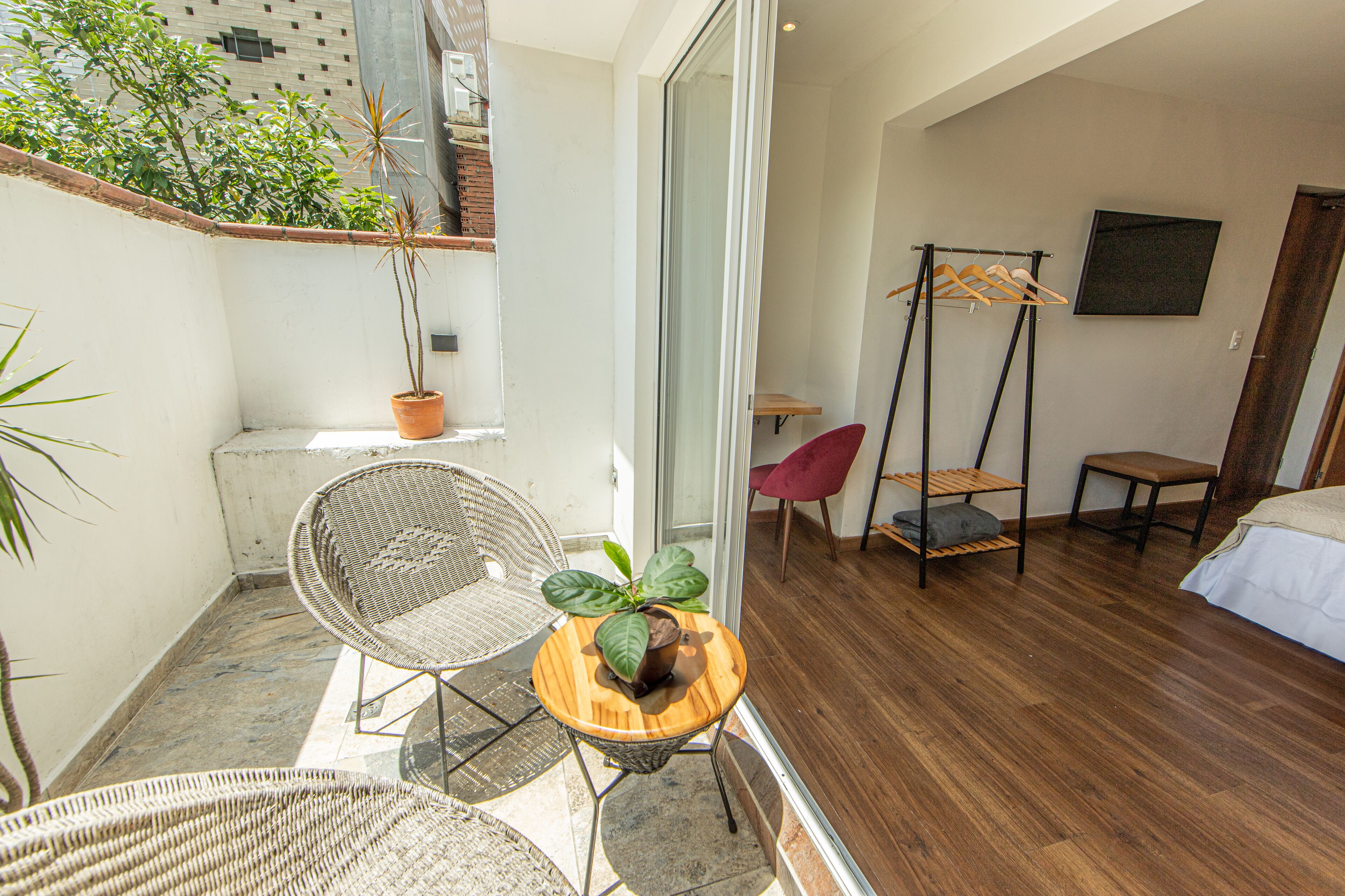 A small balcony with a wicker chair, a wooden table with a plant, a hanging chair, and a bed with a white bedspread.