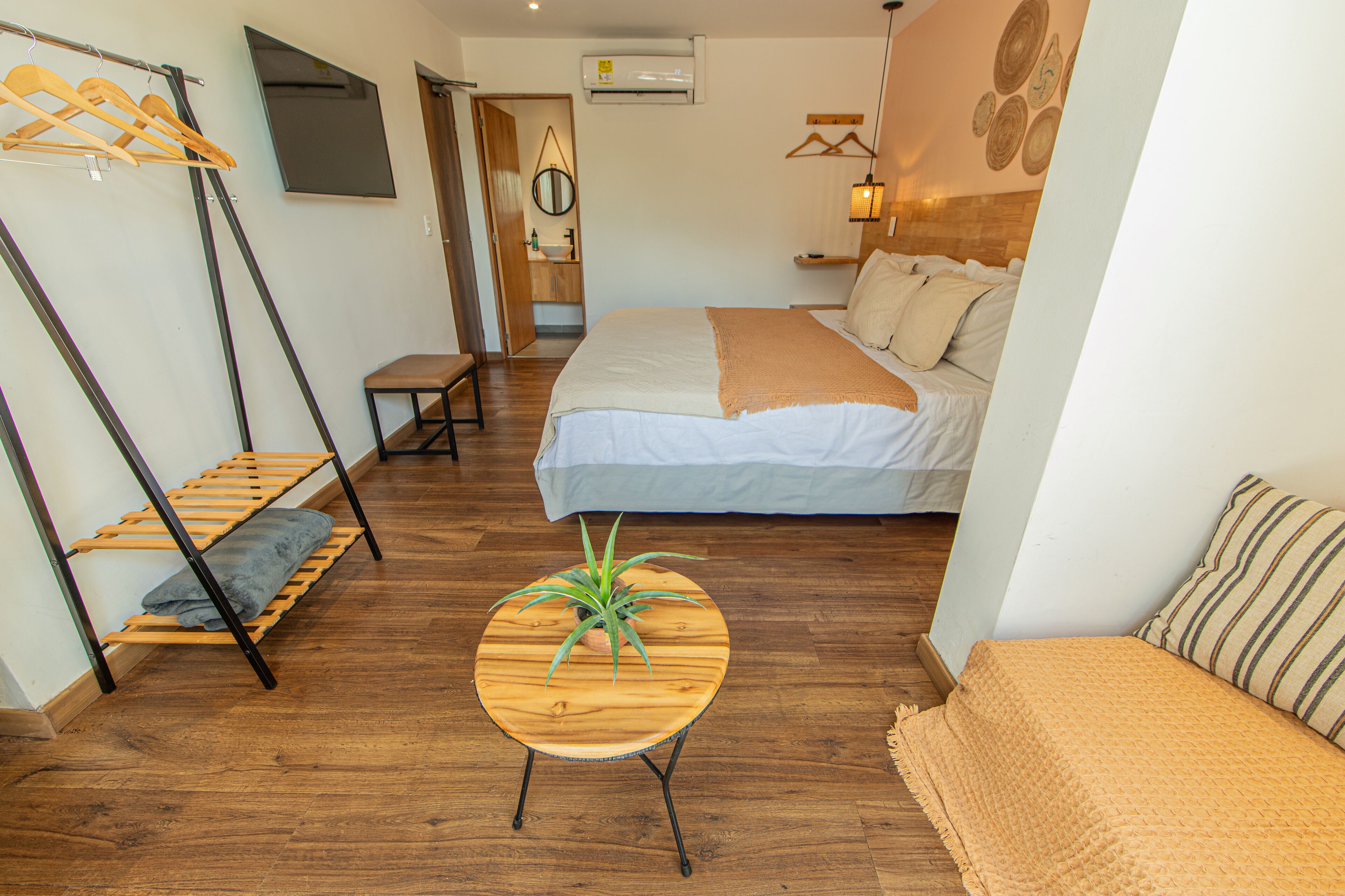 A bedroom with a bed, a wooden table with a plant, a bench, and a hanging clothes rack.