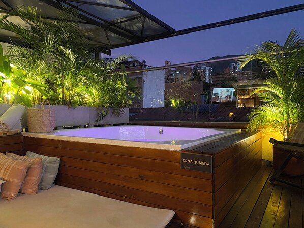 A wooden deck with a hot tub labeled 'Zona Humeda' and a view of a cityscape at dusk.