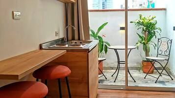 Cocina compacta con encimera de madera, pileta y dos taburetes. Posee una puerta corredera de vidrio que da a un espacio exterior con plantas en macetas y una silla.