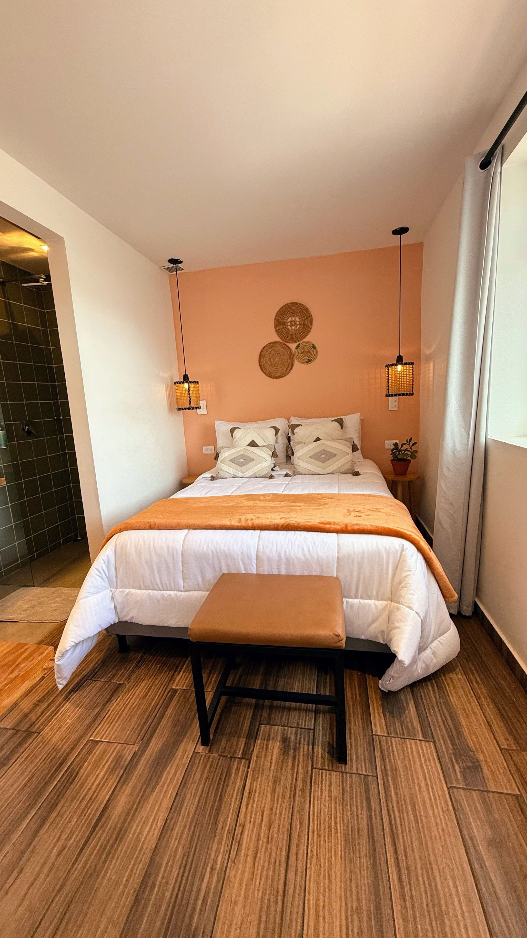 A bedroom with a bed, bedside tables, a bench, a potted plant, and a shower area.
