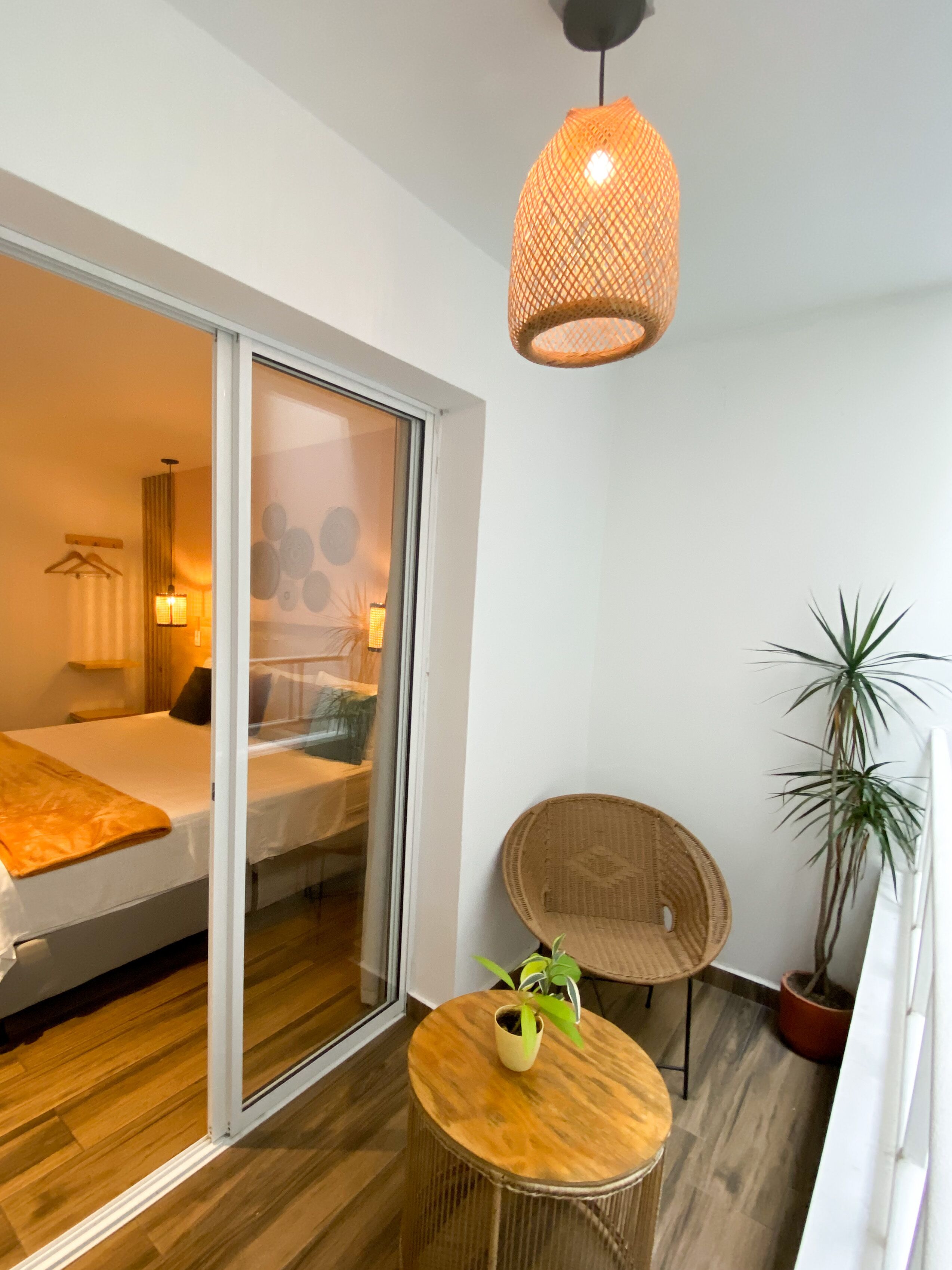 A modern hotel room with a sliding glass door leading to a balcony, a round wooden table with a potted plant, a wicker chair, and a hanging light fixture.