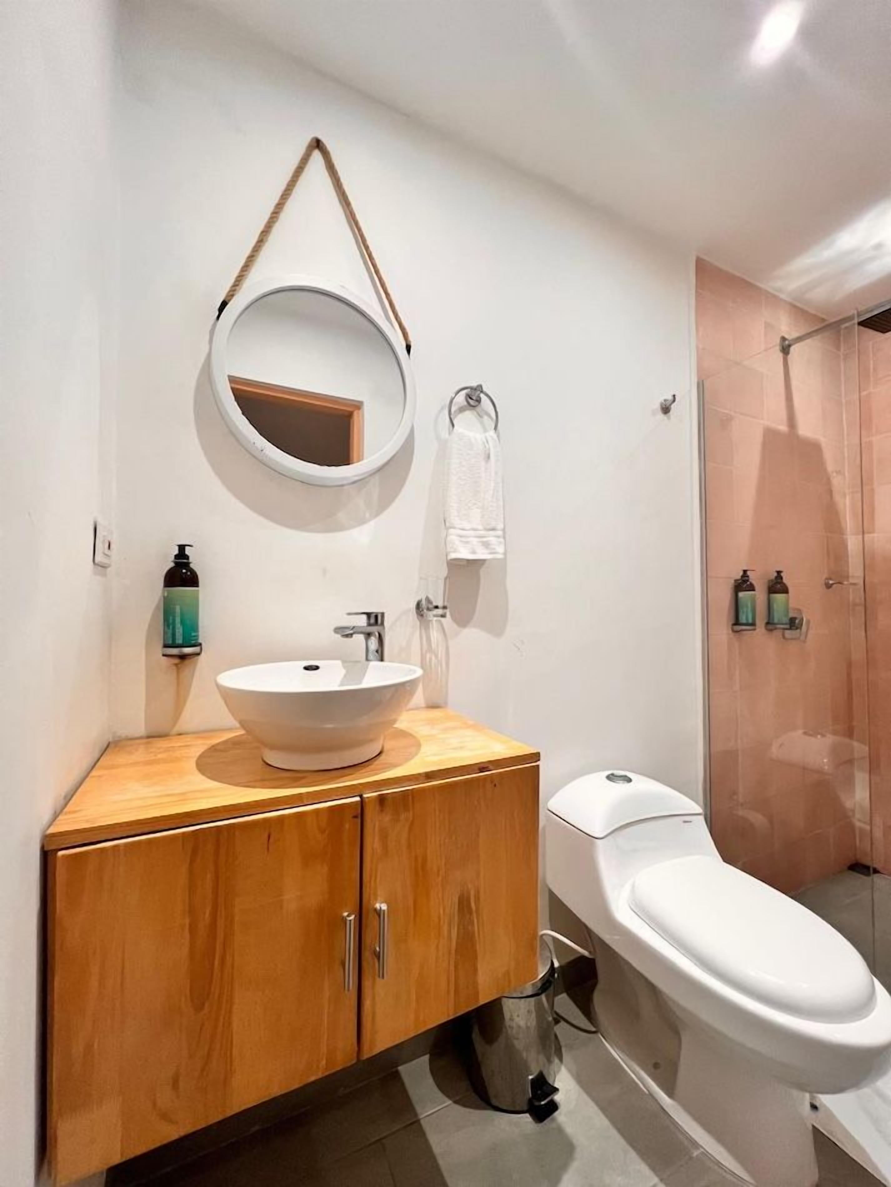 A bathroom with a wooden vanity, a white sink, a round mirror, a toilet, and a glass shower enclosure.