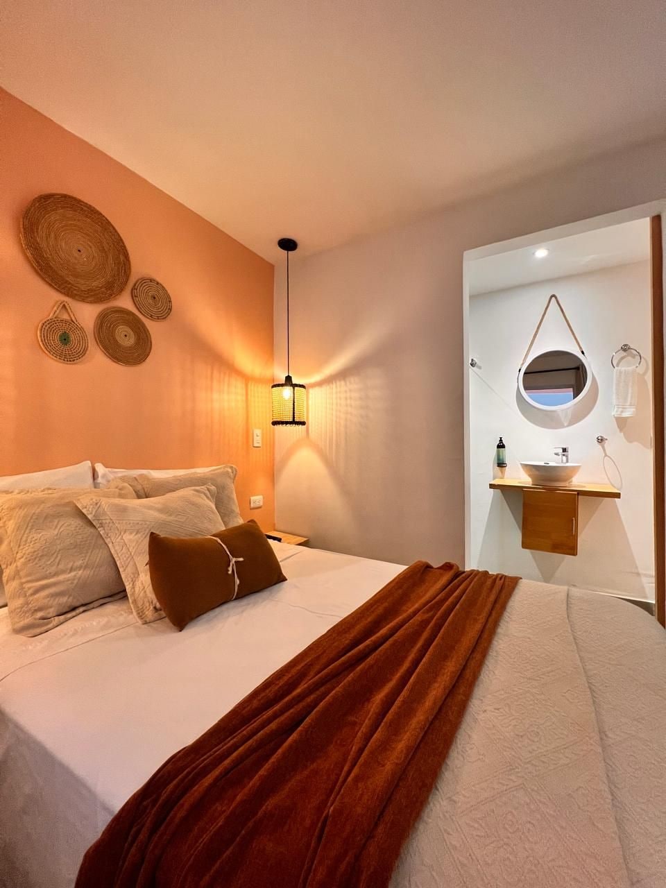 A hotel room with a bed, a bedside lamp, a round mirror, and a small shelf with toiletries.