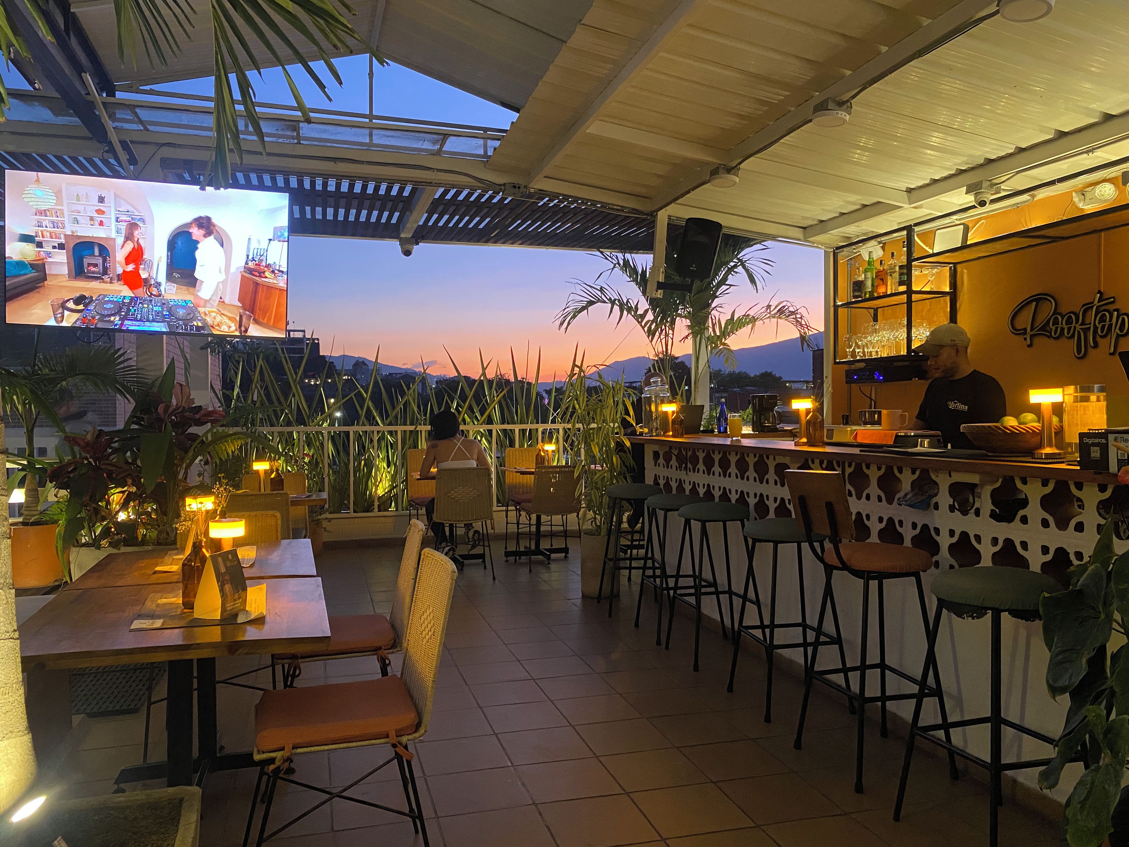 Un área de bar al aire libre con asientos, una barra y una pantalla de televisión que muestra a un DJ.