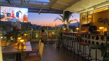 Un área de bar al aire libre con asientos, una barra y una pantalla de televisión que muestra a un DJ.