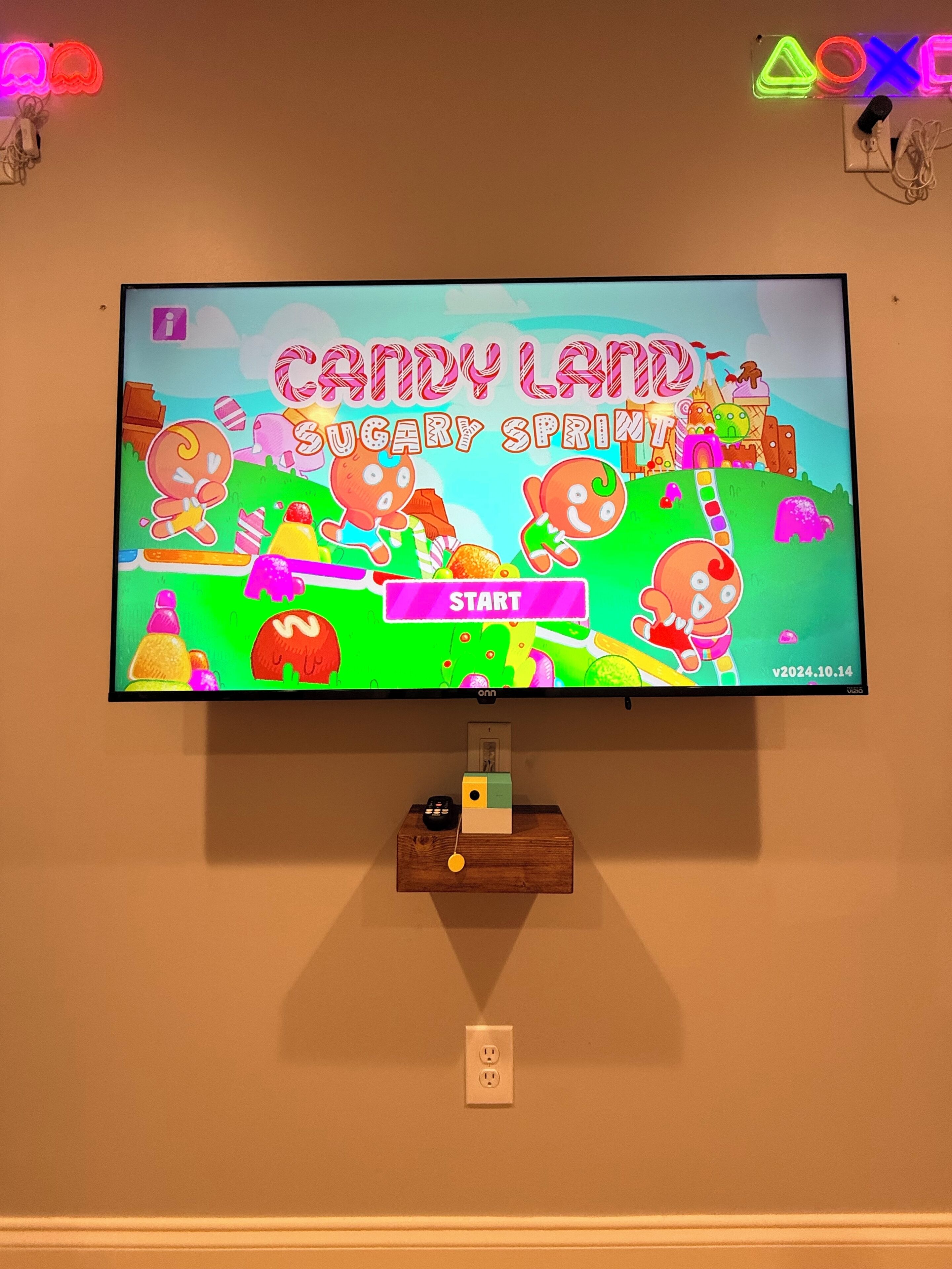 A television displaying a game titled "Candy Land Sugary Sprint" with a start button, a wooden shelf below, and a wall socket.