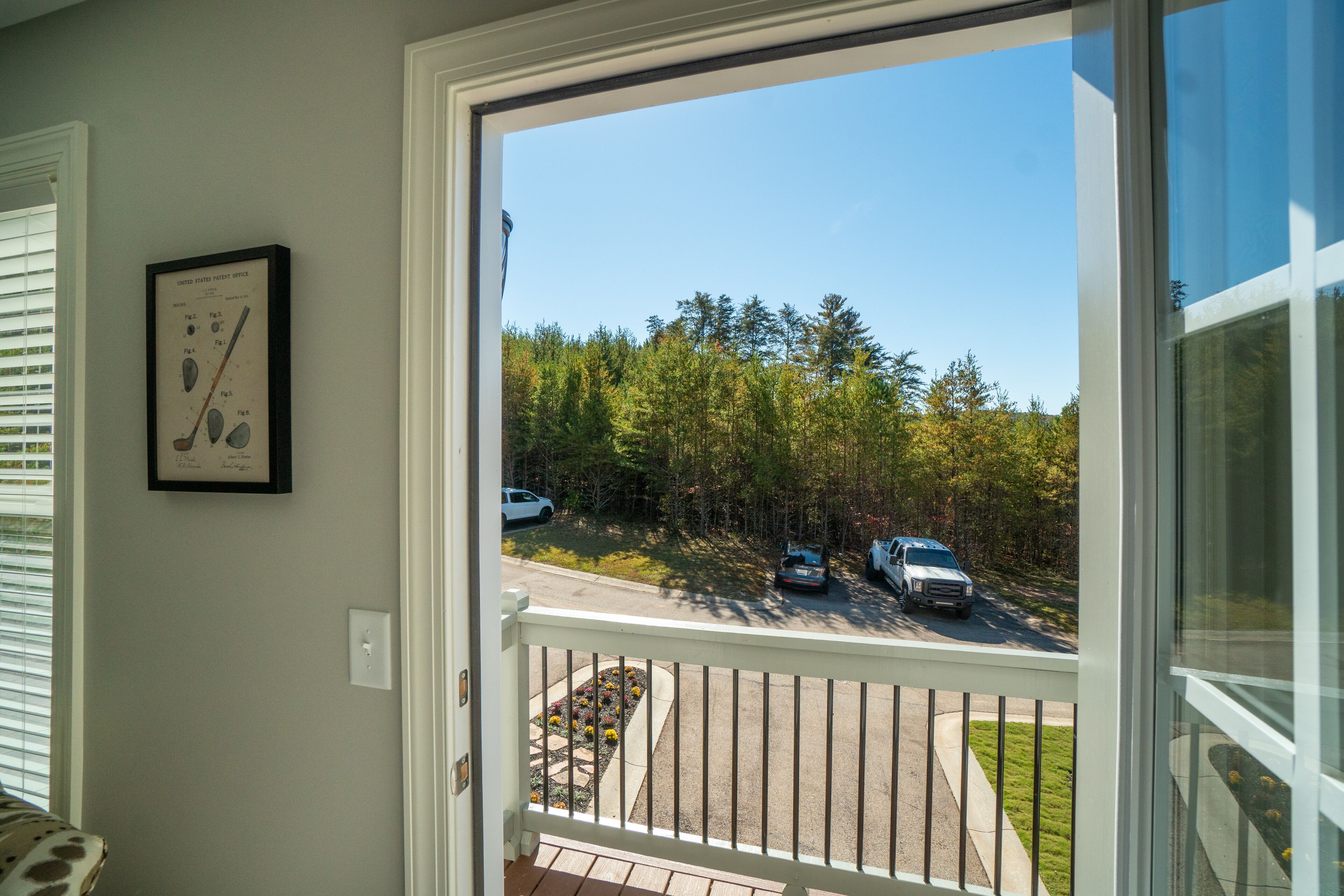 Udsigt fra en balkon med hvid gelænder, et indrammet golfscorekort på væggen, og en indkørsel med parkerede biler og en græsplæne.