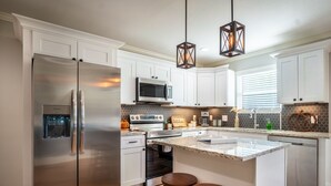 A modern kitchen with white cabinets, stainless steel appliances, a large island with stools, and decorative lighting. - Hot tub-Interactive family gaming-Game room! (Helen)