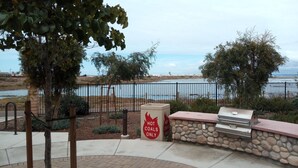 A barbecue area with a stone wall and grill, a sign indicating hot coals only, and a view of a body of water with a fence in the background. - LOWEST PRICE OF THE SEASON! 
NEW and AMAZING! Spacious! DOD, Military Welcome! (Imperial Beach)