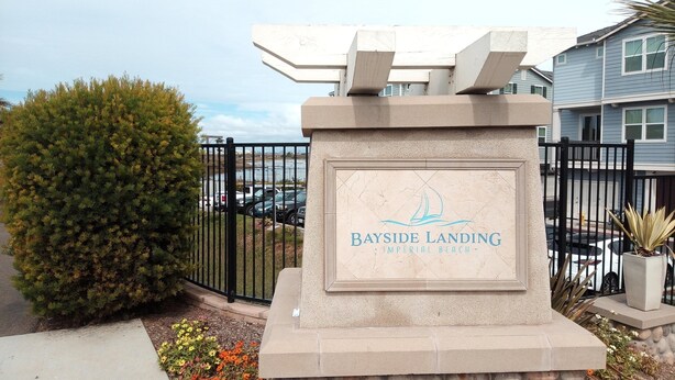 A sign for Bayside Landing Imperial Beach with a clear view of the surrounding area including a fence, parked cars, and a small tree. - LOWEST PRICE OF THE SEASON! 
NEW and AMAZING! Spacious! DOD, Military Welcome! (Imperial Beach)