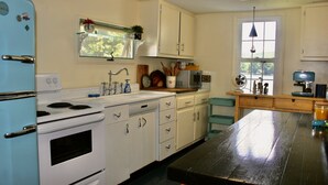 A kitchen with white cabinets, a blue refrigerator, a wooden table, and a window with a view of trees. - Cozy cottage perfect for family and friends (Hamilton)