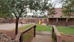 A wooden deck leading to a backyard with a play area, including a slide and swings, and a building with a red-tiled roof. - Family Fun "Campout" (Saint George)
