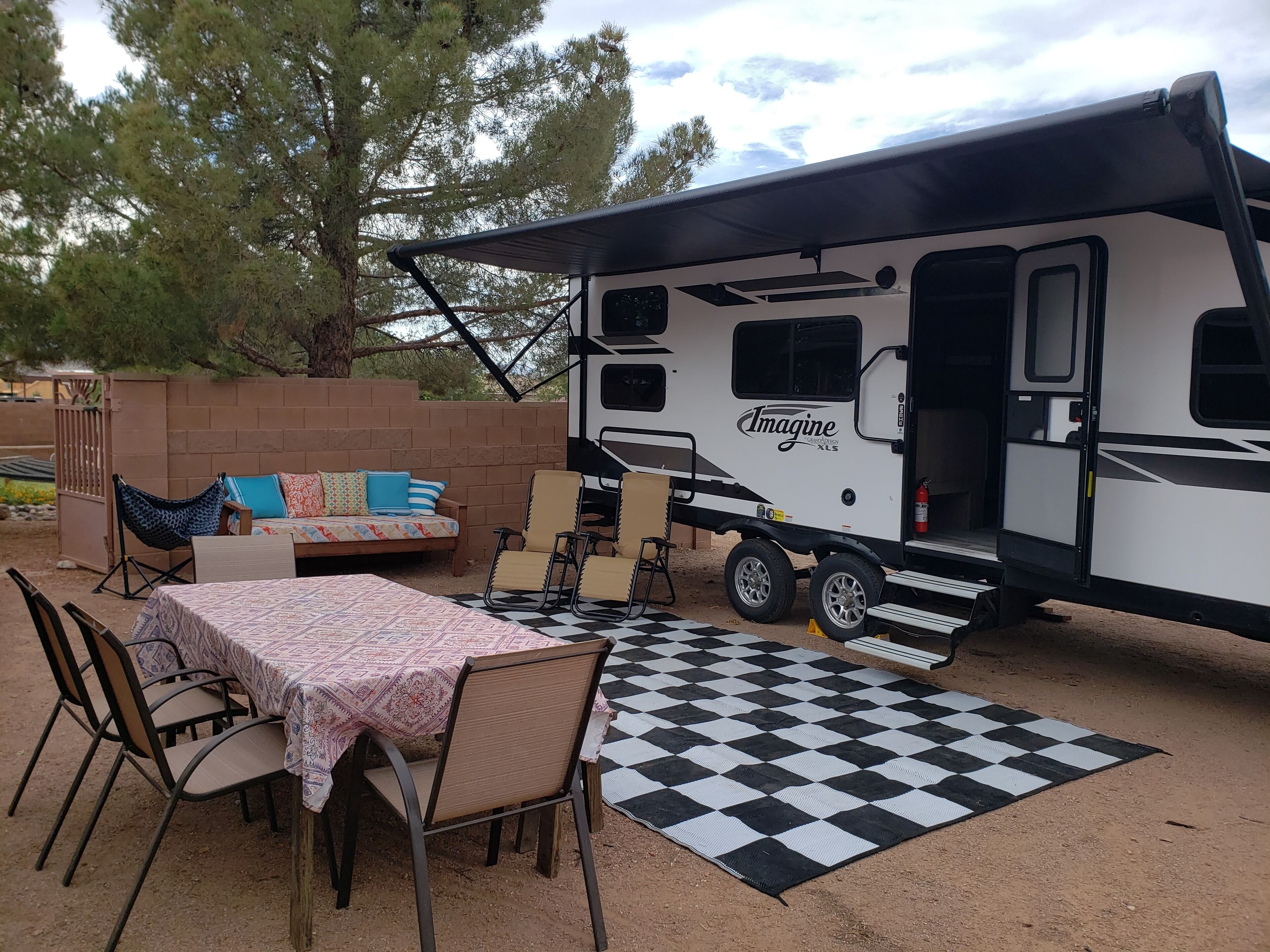 An RV with a covered porch, a dining table with chairs, and a checkered outdoor rug.