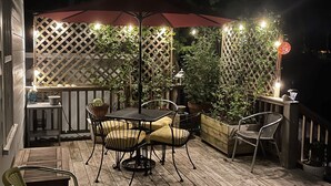 Een terras buiten met een houten terras, een tafel met stoelen, een rode parasol en lichtslingers.