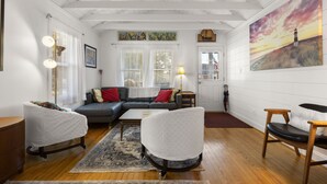 Een gezellige woonkamer met een hoekbank, twee fauteuils, een salontafel en een vloerkleed. Op de muur hangt een schilderij van een vuurtoren, en er is een deur met een ruit.