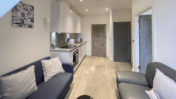 A modern apartment with a kitchenette, a dark grey sofa, a light wooden floor, and a door leading to another room. - Remarkable 1-Bed Apartment in Slough (Slough)