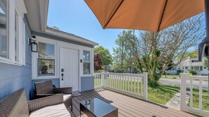 Une terrasse dotée de meubles en osier et d’une table en verre, offrant une vue sur une clôture blanche et une rue.