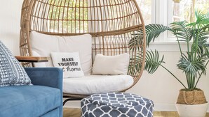 Une chaise en osier avec un coussin, un canapé bleu, un pouf à motifs et une plante en pot.