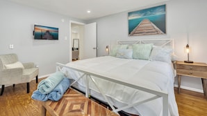 Une chambre à coucher avec un lit, une chaise, un plancher en bois, une table de chevet et un tableau accroché au mur.