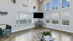 Een ruime woonkamer met een groot raam, een flatscreen-tv, een glazen salontafel met bloemen en een houten plafond.