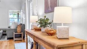 Une console en bois, agrémentée de deux lampes, d’une plante en pot et de paniers tressés, se trouve dans un salon lumineux, où l’on aperçoit un canapé gris et une fenêtre en arrière-plan.