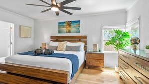 Une chambre avec un lit en bois, un ventilateur de plafond, une table de chevet, une commode, une fenêtre donnant sur un beau paysage et un tableau accroché au mur.