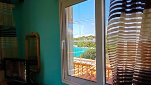 A room with a window overlooking a coastal area, a wooden nightstand with a microwave, and striped curtains. - Iliaktis Xenios Zeus  (Νήσοι)