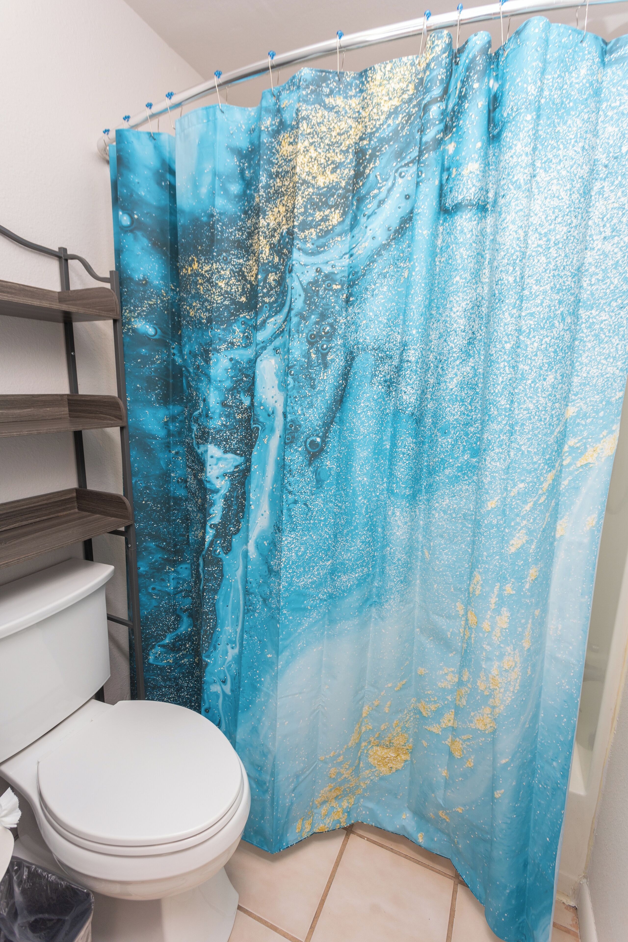 A bathroom with a white toilet, a blue shower curtain with gold splatter, and a tiled floor.
