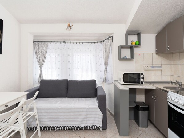A compact kitchenette with a microwave, stove, and sink. A gray sofa with a striped blanket is positioned in front of a window with sheer curtains. - Apartment Mia by Interhome (Novigrad (Zadar))