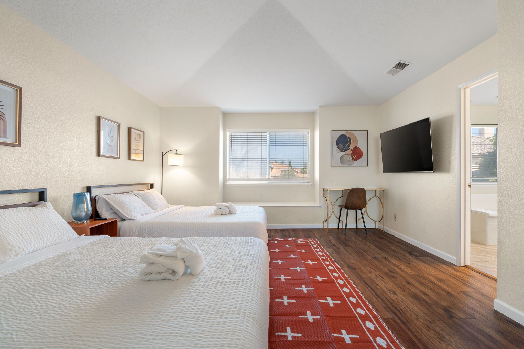 Habitación de hotel con dos camas, un televisor, una ventana con persianas y una alfombra roja estampada.