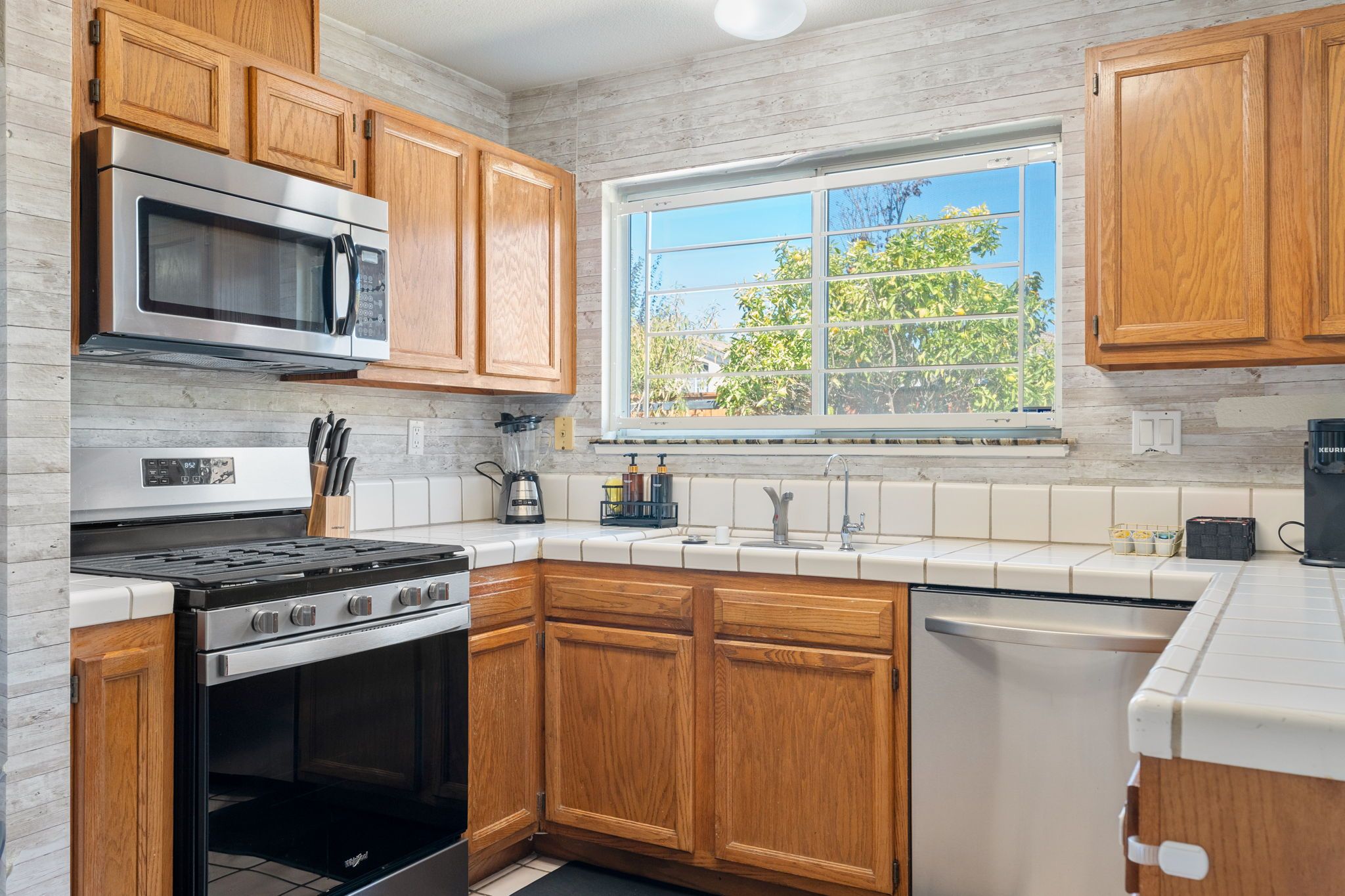 A kitchen with wooden cabinets, a stainless steel stove, a microwave, and a window with a view of trees.