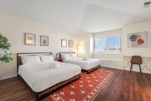 A hotel room with two beds, a red patterned rug, a desk with a chair, and artwork on the walls.