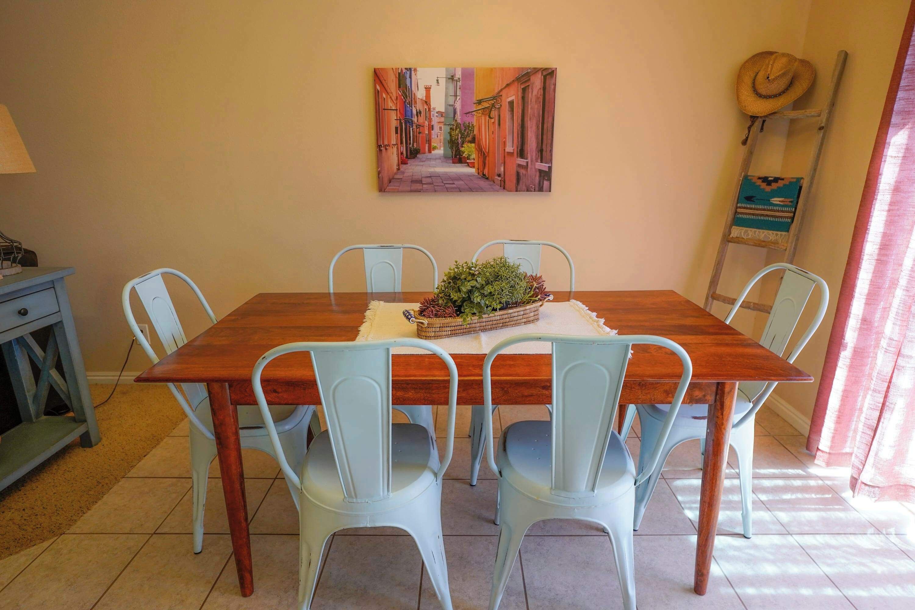 A dining area with a wooden table, four light blue chairs, a potted plant, and a painting of a street scene on the wall.