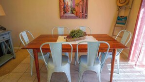 A dining area with a wooden table, four light blue chairs, a potted plant, and a painting of a street scene on the wall.