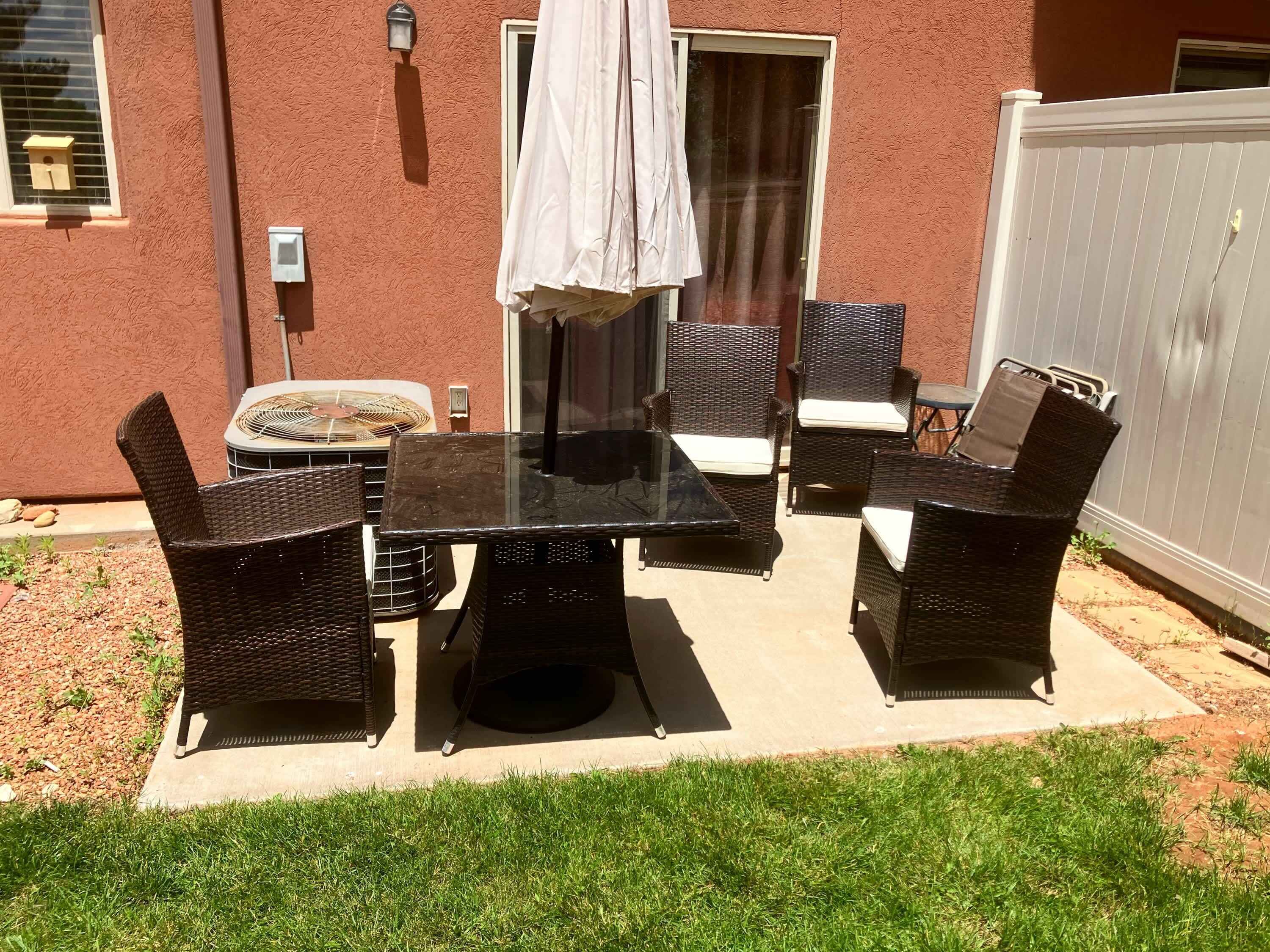An outdoor patio with a glass table and wicker chairs, a white umbrella, and a small air conditioning unit.