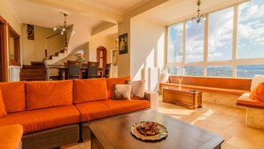 A spacious living room with a large orange sofa, a wooden coffee table, and a view of the sea through large windows.