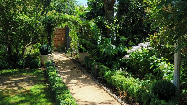 Un sendero en un jardín bordeado por setos y árboles bien cuidados, que conduce a un portón de madera.