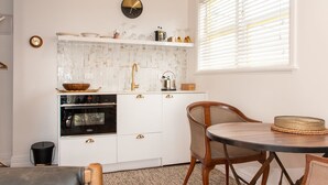 Una cocina con armarios blancos, un horno negro, una mesa de comedor de madera y una ventana con persianas.