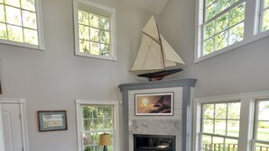 A spacious living room with a fireplace, large windows, and a sailboat decoration. - Beautiful Modern Cottage with Private Lake Access (Skaneateles)