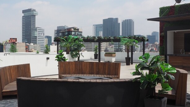 A rooftop terrace with wooden seating, potted plants, and a view of city buildings. - Loft Sinaloa By The Local Way (Mexico City)
