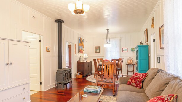 A living room with a sofa, wooden coffee table, and a fireplace. There is a dining area with a table and chairs, and a kitchen area with cabinets and a stove. - Historic Turn-of-the-Century Bungalow in the heart of Volcano Village. (Volcano)