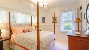 A four-poster bed with a floral bedspread, a bedside table with a lamp, and a window with curtains. - Historic Turn-of-the-Century Bungalow in the heart of Volcano Village. (Volcano)