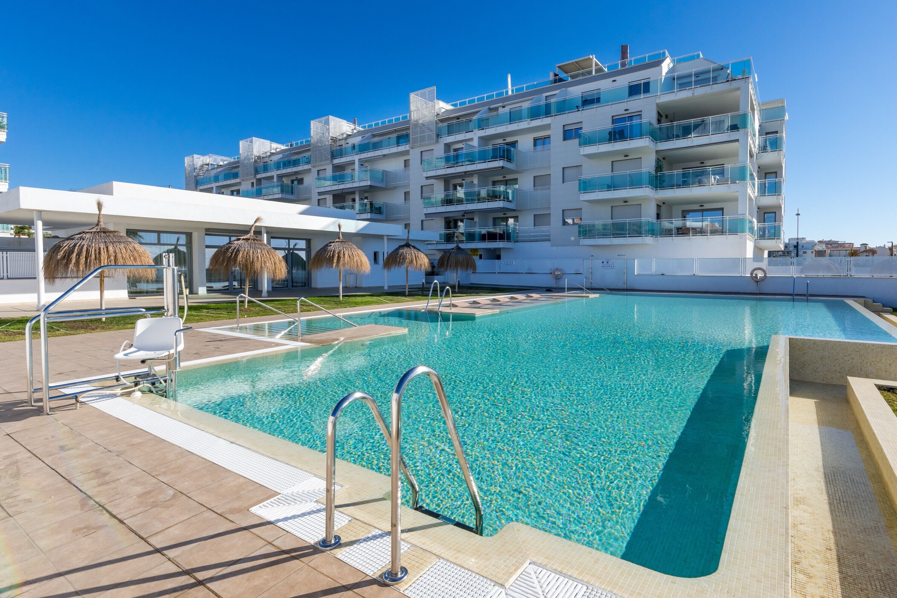 A swimming pool with a clear blue water, surrounded by a white building with balconies and a covered area with chairs.