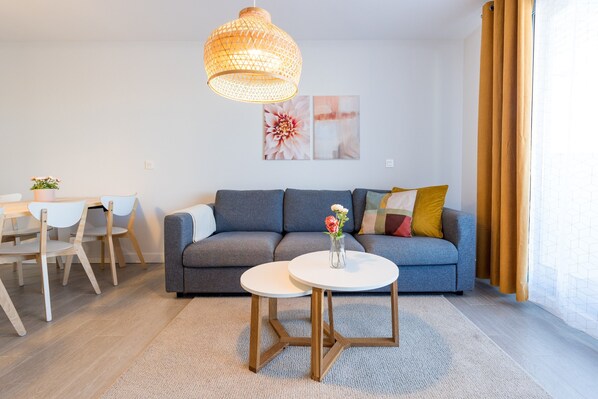 A modern living room with a grey sofa, two round tables, a hanging woven light fixture, and a dining area with a white table and chairs.
