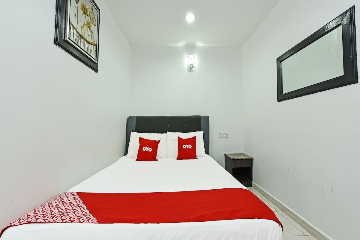 Ein Schlafzimmer mit einem Bett, zwei Kissen, einem Nachttisch und einem Spiegel an der Wand. Eine Deckenleuchte ist ebenfalls vorhanden.