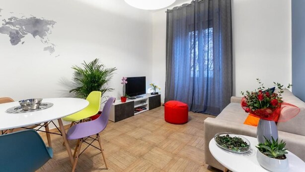 A modern living room with a round dining table, colorful chairs, a red ottoman, a sofa, a TV, and a world map on the wall.